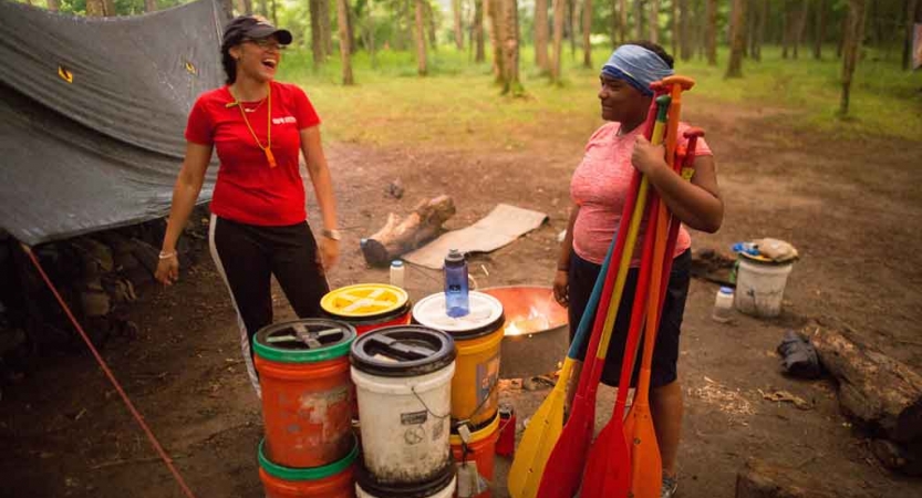 Two people stand at a campsite in a wooded area. One of them holds several paddles. 